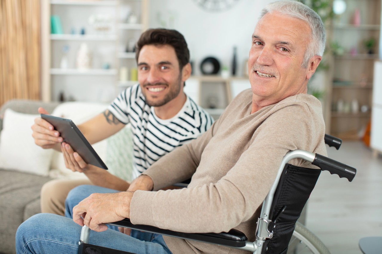 Elderly man in wheelchair smiling while his support worker shows him information on a tablet device at home