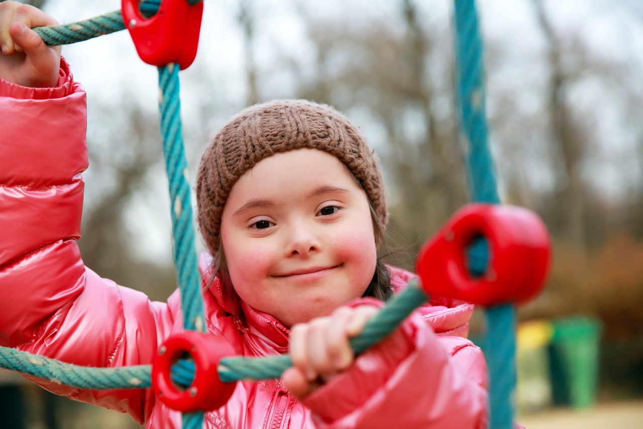 Smiling young girl with Down syndrome playing on climbing equipment at a playground, demonstrating inclusion and community participation