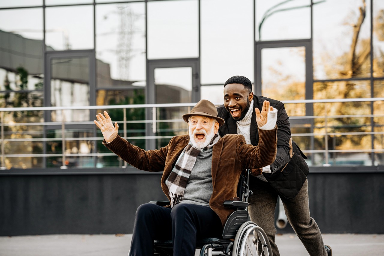 Joyful elderly man in wheelchair with arms outstretched alongside his disability support worker outdoors