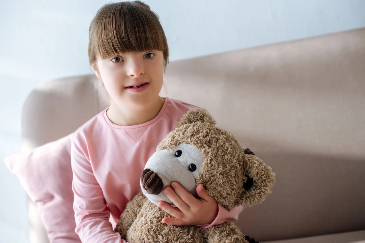 Young girl with Down syndrome sitting on a couch holding a teddy bear, representing the people disability support workers assist