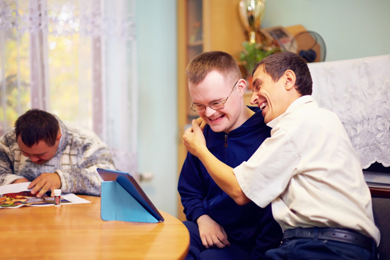 Young men with intellectual disabilities laughing together while using a tablet device in a supported living environment